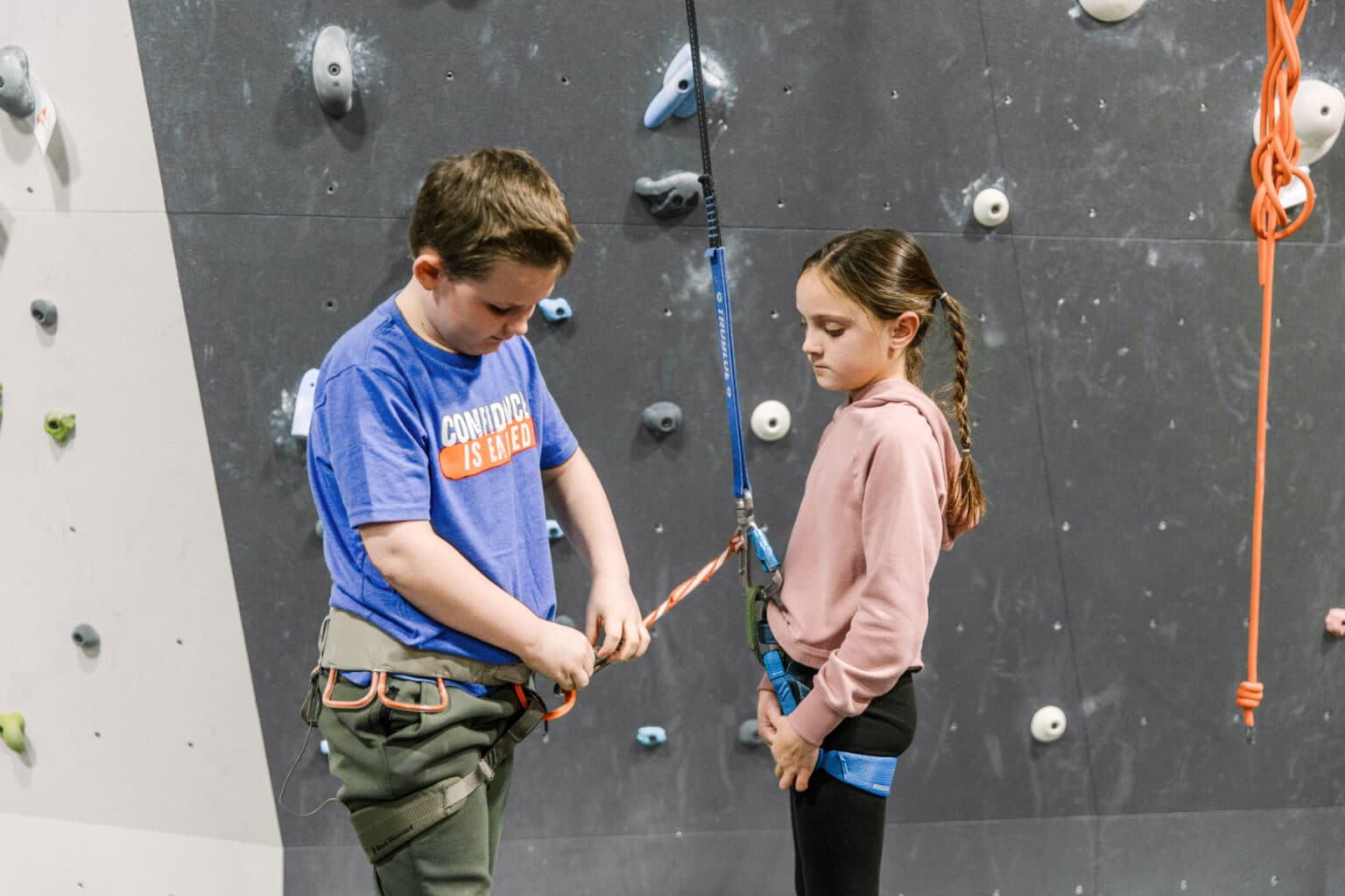 Two children are positioned on a climbing wall indoors, engaging with an auto belay for secure climbing.