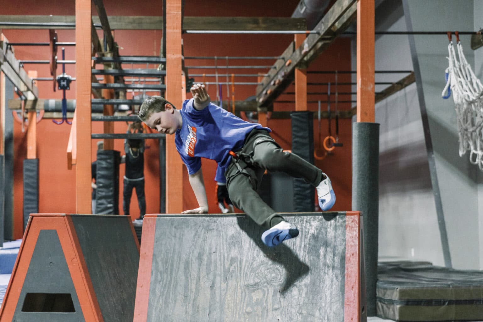 A young person in a blue shirt and black pants is climbing over a slanted obstacle in an indoor gym with red walls and various equipment.