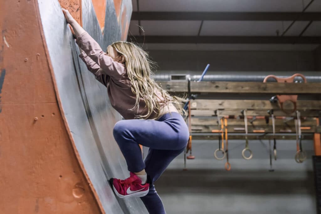 A woman ascends a warped wall in an indoor gym, showcasing her strength and determination in a vibrant environment.